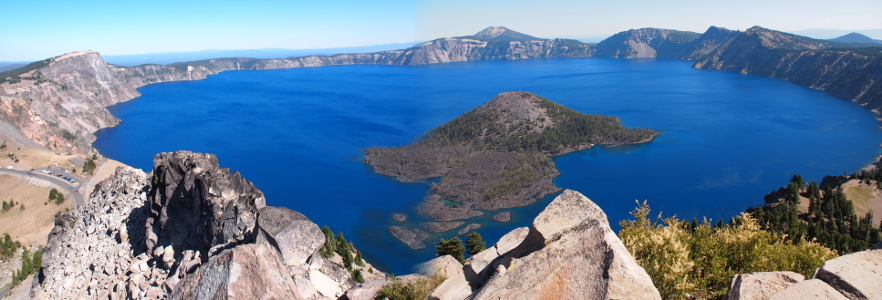 [Deep, deep blue lake ringed by crater rock walls with a treed island in it.]